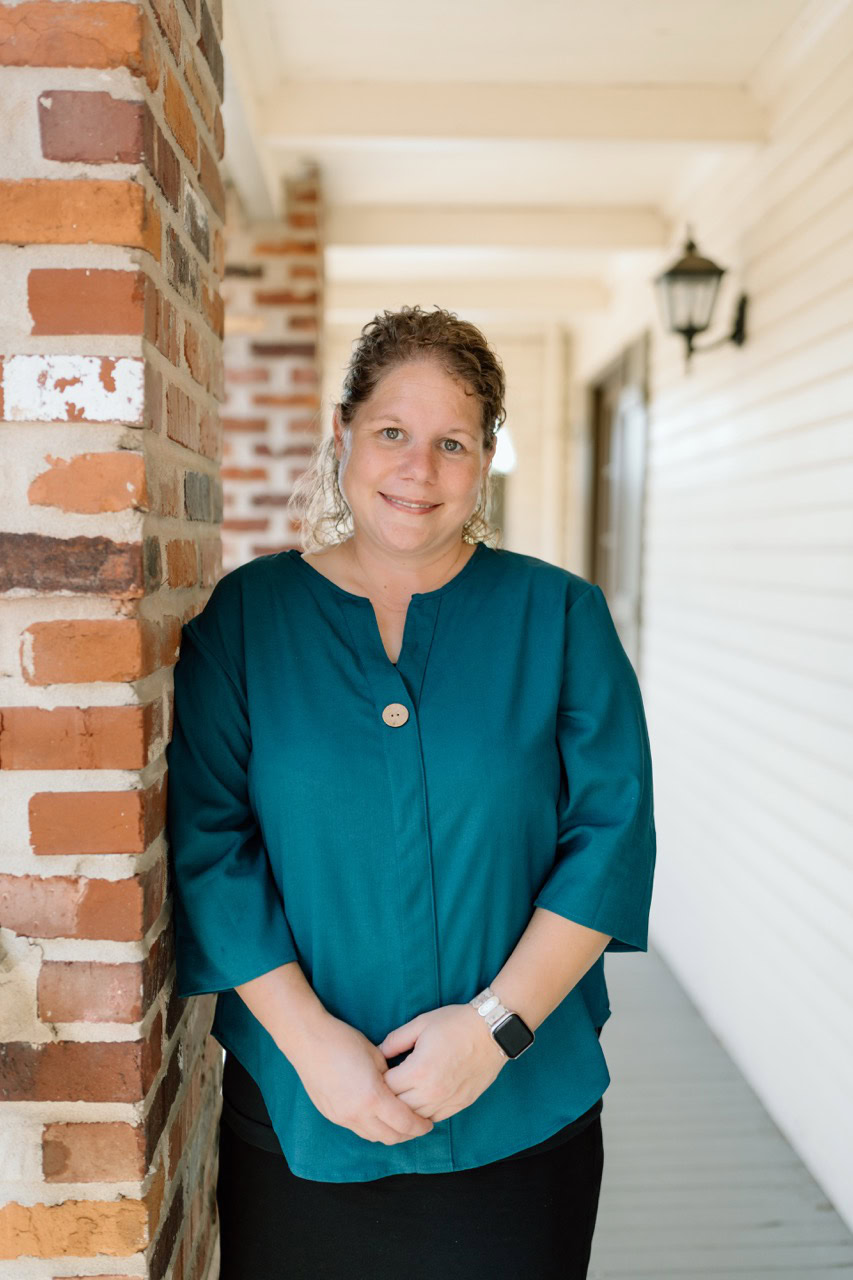 A friendly woman in a teal blouse standing on a porch with brick and white siding, smiling at the camera.