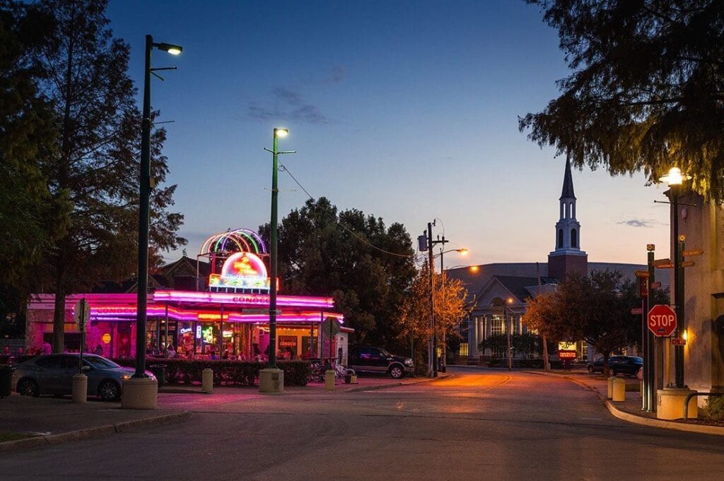 Brightly lit diner with neon signs and a church steeple in a small town setting, highlighting local community, nightlife, and Southern charm.