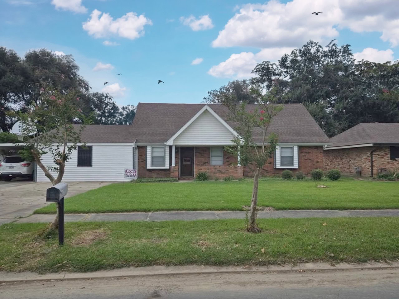 A welcoming suburban home featuring a well-maintained front lawn, mature trees, and a clear blue sky. The house has a brick and siding exterior, with a driveway and mailbox visible.